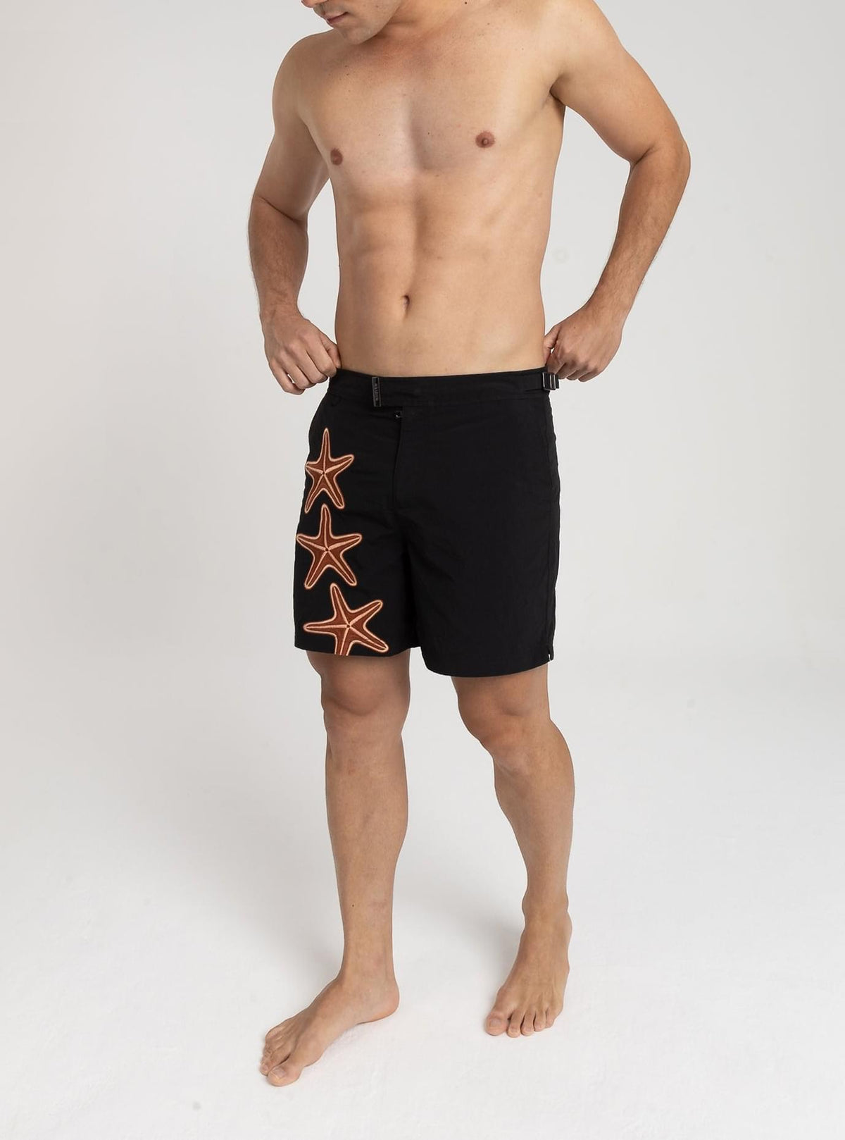 Modelo usando o short de praia RAUTER TRÊS ESTRELAS MÉDIO, bordado industrial de estrelas-do-mar sobre nylon reciclado, corte médio.